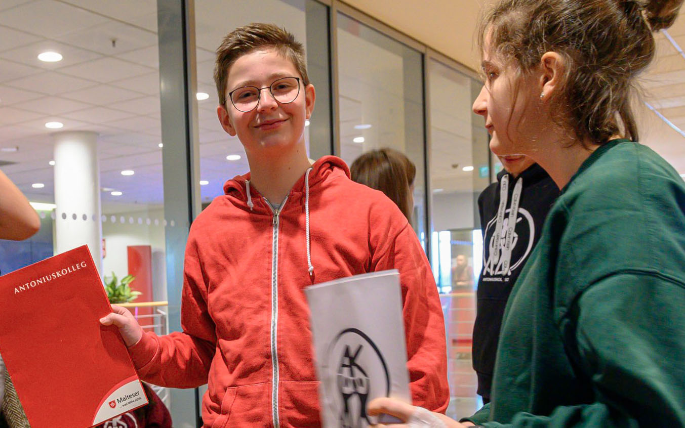 Junge mit rotem Pullover lächelt in die Kamera und hält eine rote Mappe aus Papier in der Hand